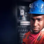 Electricista con casco y lentes de seguridad frente a un tablero eléctrico
